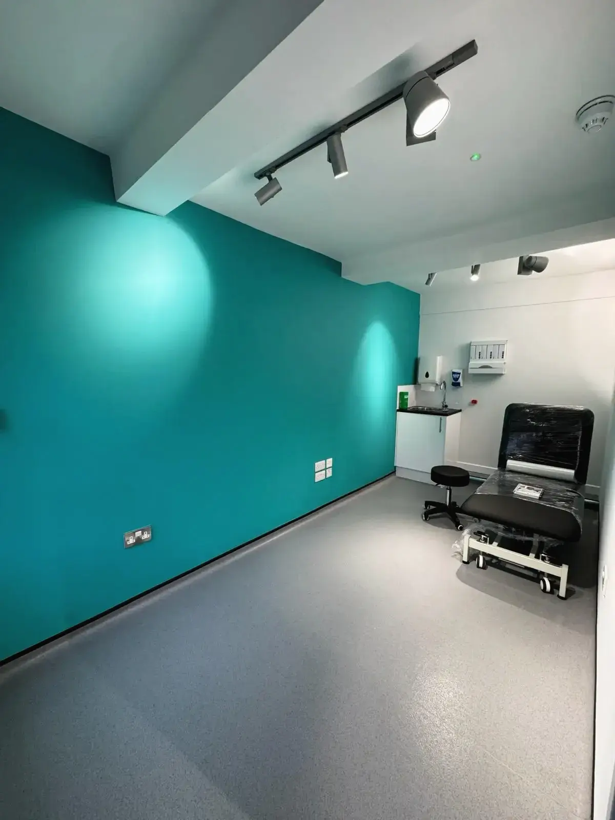 Clean consultation room with teal accent wall, medical bed, and spot lighting