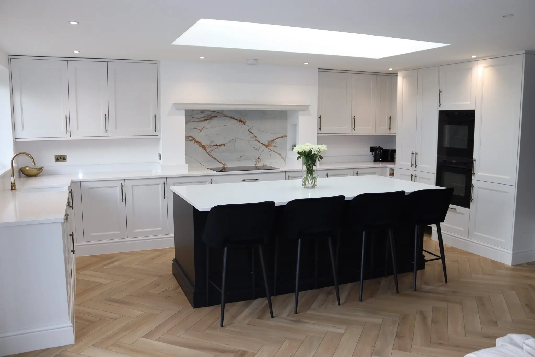Luxury open-plan kitchen renovation featuring white shaker cabinets, a dark kitchen island, herringbone flooring, brass fixtures and a skylight.